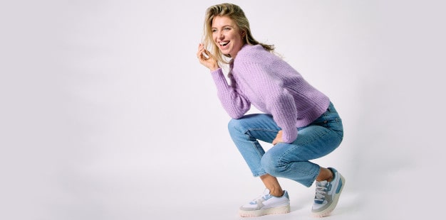Lachende Frau vor weißem Hintergrund. Sie trägt Pullover, Jeans und Puma Cali Dream Pastel Wns Sneaker in Weiß